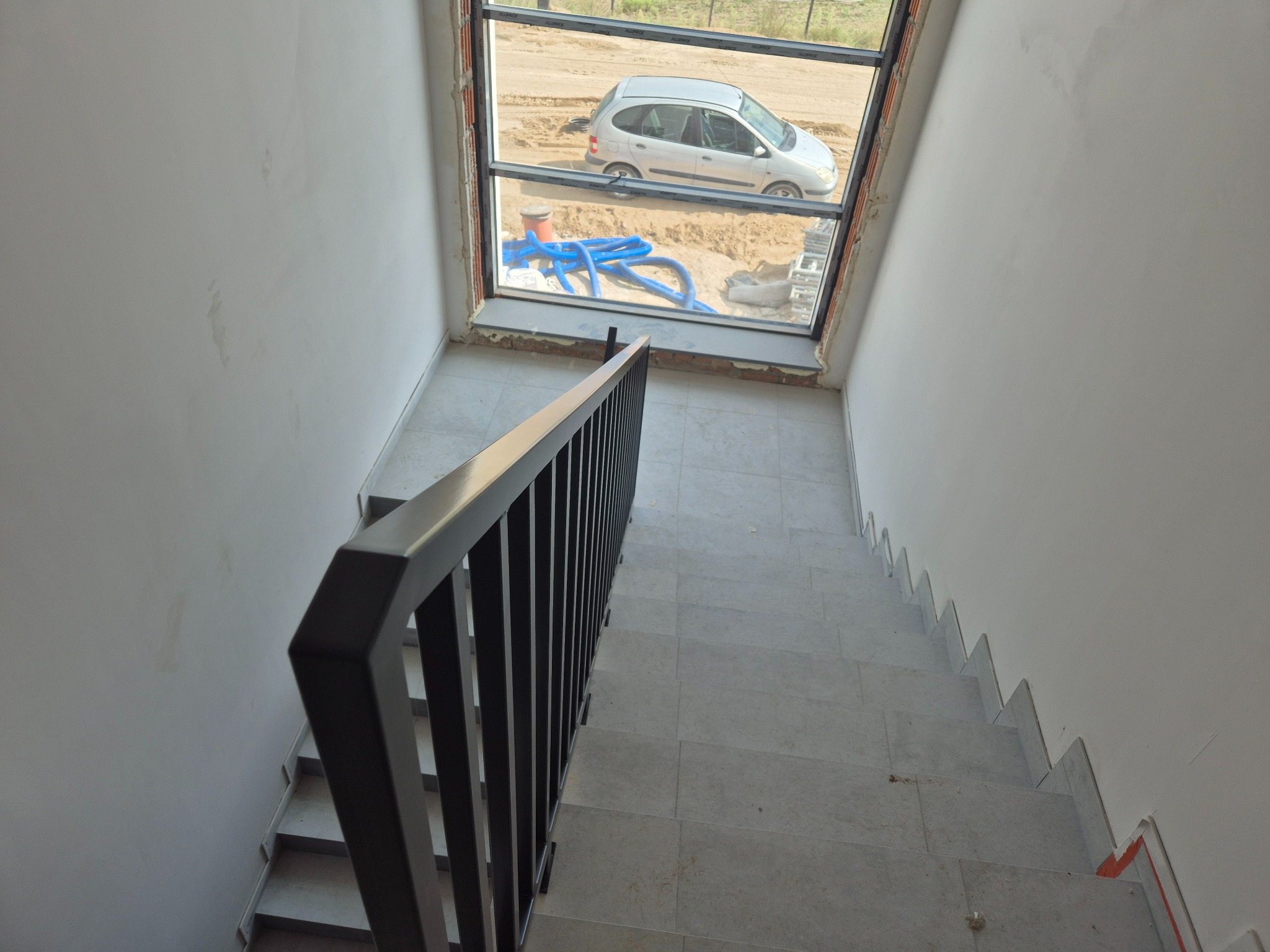Wewnętrzne schody z czarną metalową balustradą, widok z góry na dół, w tle okno z widokiem na plac budowy i zaparkowany samochód.