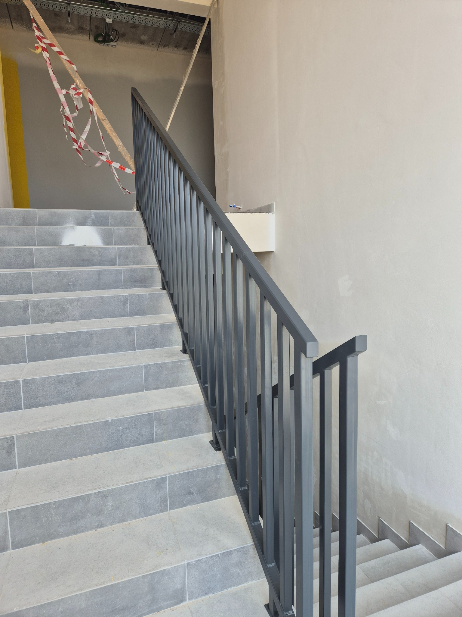 Szare schody wyłożone płytkami z metalową balustradą o pionowych szczeblach, widoczne taśmy ostrzegawcze i surowy stan wykończenia wnętrza.
