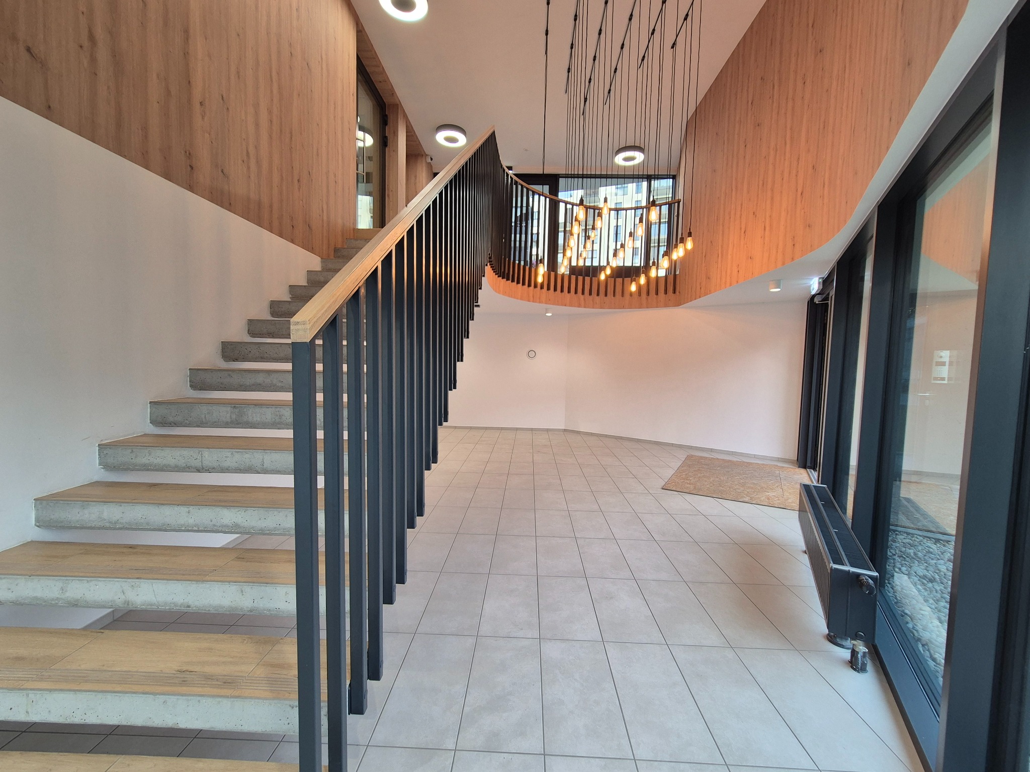 Wnętrze nowoczesnego budynku z betonowymi schodami z drewnianymi stopniami i czarną balustradą, oświetlone wiszącą lampą z żarówkami w stylu retro.