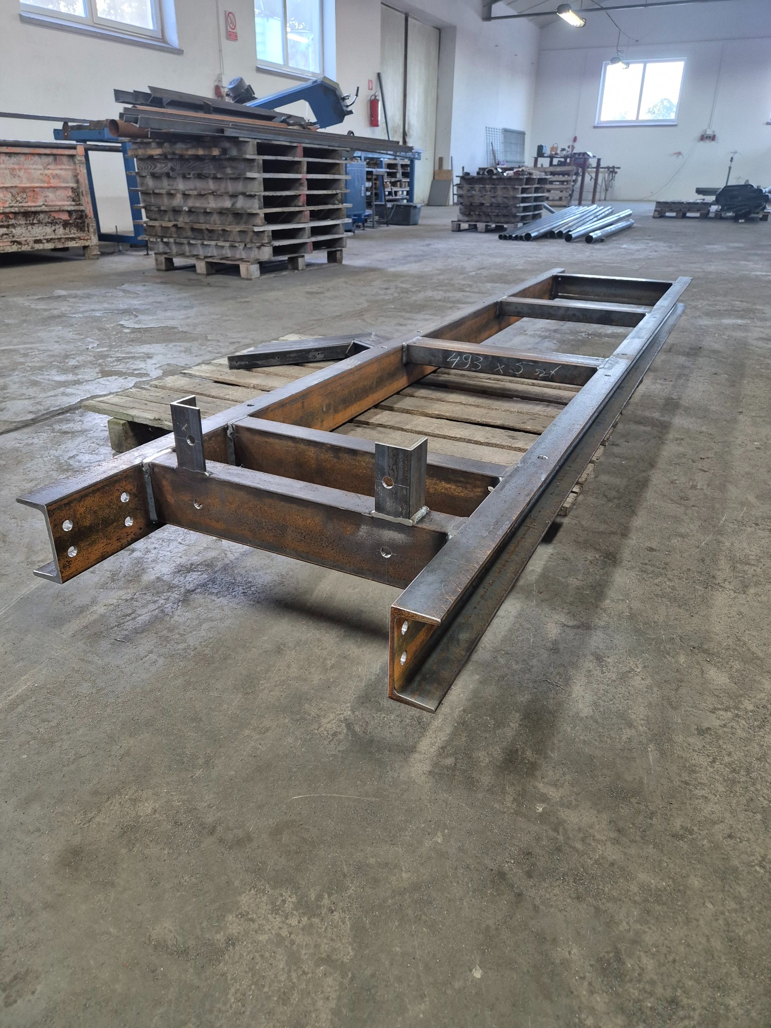 Metalowa rama konstrukcyjna z profili stalowych na palecie w hali produkcyjnej, widoczne spawane łączenia i otwory montażowe, w tle inne konstrukcje i materiały.