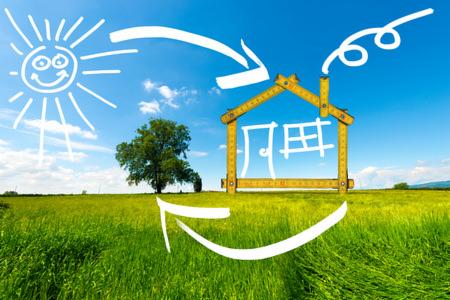 Stylizowany dom ułożony z żółtej miarki stolarskiej na tle zielonej łąki, błękitnego nieba i narysowanego słońca z uśmiechem, z dodatkowymi strzałkami symbolizującymi ruch.