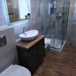 Nowoczesna łazienka z kabiną prysznicową, umywalką nablatową na szafce z ciemnego drewna, podświetlanym lustrem LED i brązowymi płytkami imitującymi drewno na podłodze.