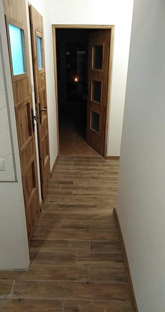 Korytarz z drewnopodobną podłogą z płytek, dwoje drewnianych drzwi z szybami, jedne otwarte, widok w głąb mieszkania.
