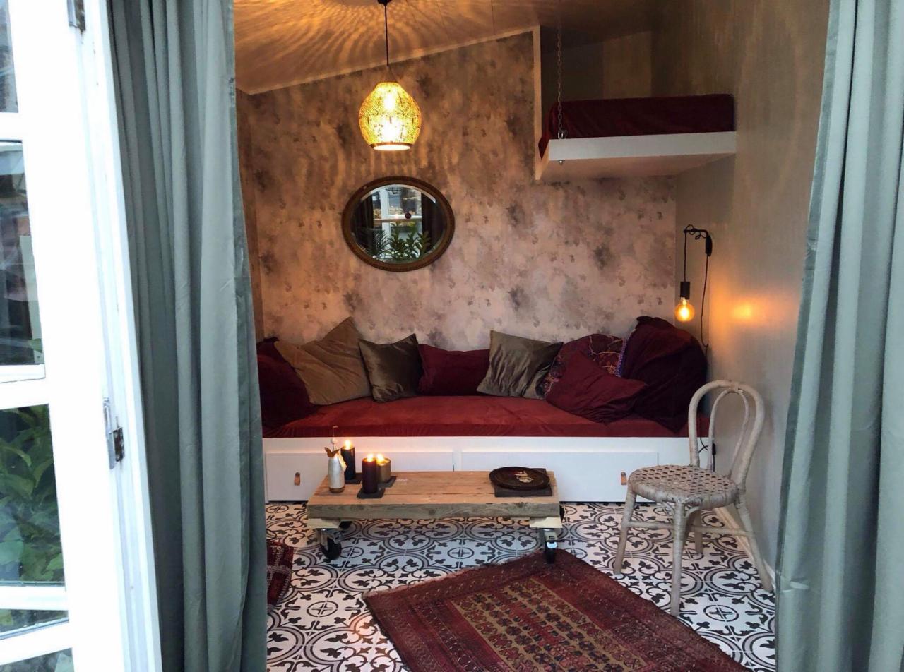 Przytulny pokój z niskim łóżkiem/sofą w kolorze burgundu, ozdobnymi poduszkami, drewnianym stolikiem kawowym na kółkach, dywanem orientalnym i wiszącym łóżkiem na antresoli, oświetlony dekoracyjną...