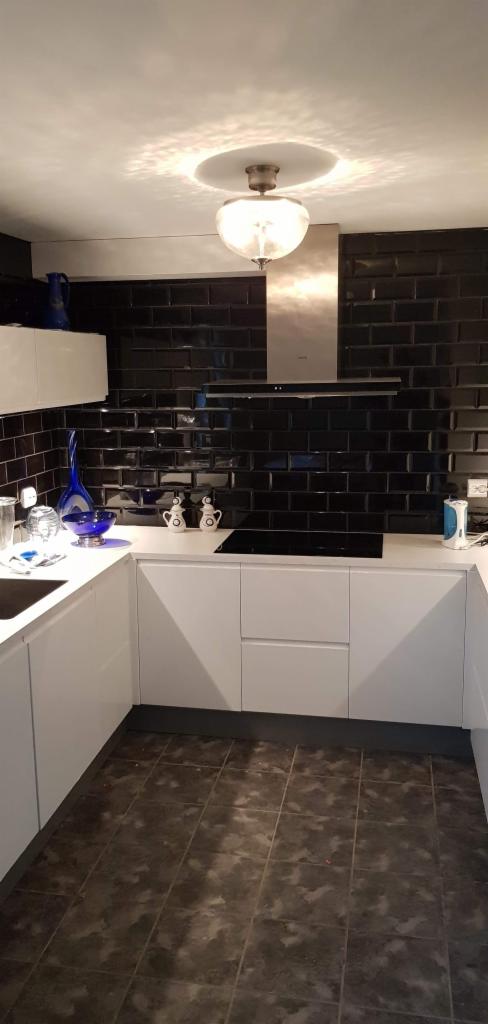 Nowoczesna kuchnia z białymi szafkami, czarną, błyszczącą mozaiką na ścianie, płytą indukcyjną i okapem ze stali nierdzewnej, oświetlona wiszącą lampą z mlecznego szkła.