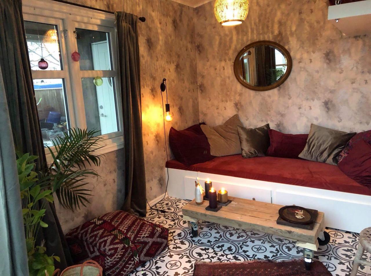 Przytulny kącik wypoczynkowy w stylu boho z niskim siedziskiem z bordową tapicerką i poduszkami, stolik kawowy na kółkach z surowego drewna, wzorzysta podłoga i ściana z efektem betonu.