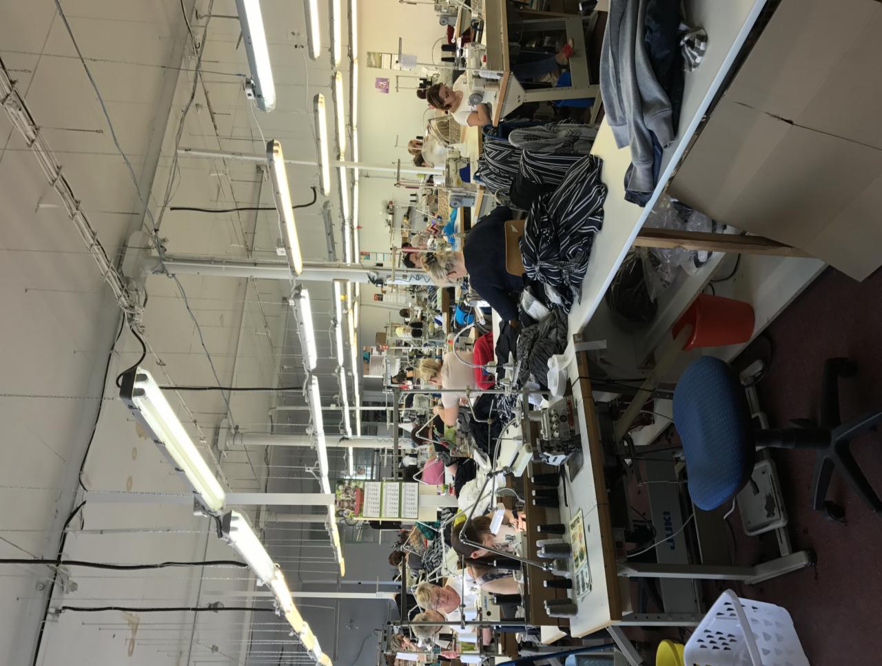 Wnętrze hali produkcyjnej z rzędami stanowisk do szycia, oświetlonych lampami jarzeniowymi, z pracownikami przy maszynach i stertami materiałów.