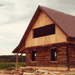Surowy stan budowy domu z bali z brązowym dachem, widoczne puste otwory okienne i sterta drewna na ziemi.