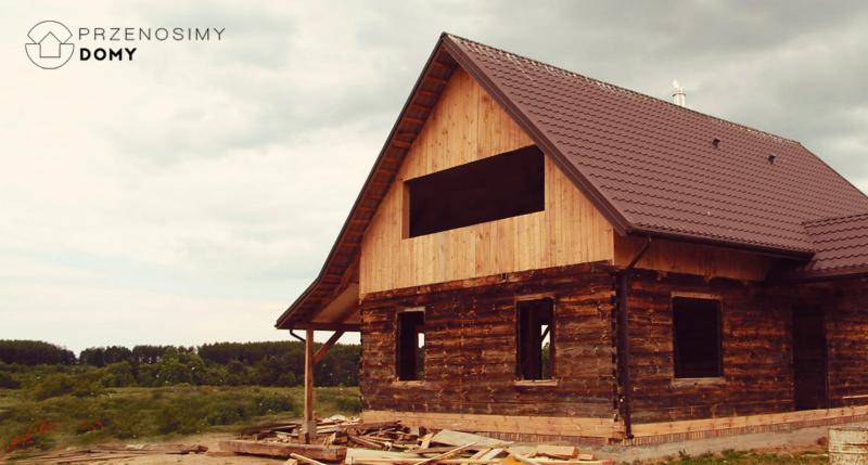 Surowy stan budowy domu z bali z brązowym dachem, widoczne puste otwory okienne i sterta drewna na ziemi.