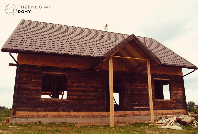 Surowy, ciemnobrązowy szkielet domu z bali z brązowym dachem, widoczne niezabudowane otwory okienne i drzwiowe, częściowo wykończony fundament, drewniane podpory ganku, logo firmy w lewym górnym rogu.