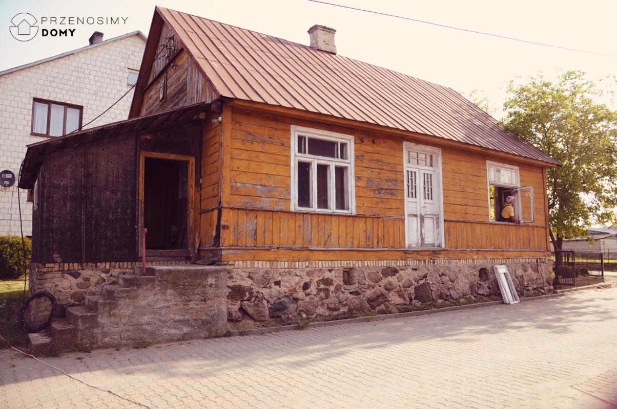 Drewniany dom z pomarańczową elewacją, widoczne ślady użytkowania, kamienna podmurówka, w oknie mężczyzna w żółtej koszulce, obok oparta o ścianę biała rama okienna.