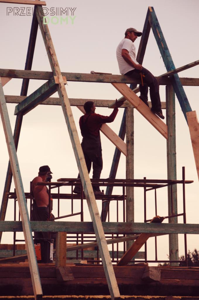 Trzech robotników pracuje przy montażu drewnianej konstrukcji domu, jeden siedzi na górnej belce z młotkiem, drugi podaje deskę, trzeci stoi na rusztowaniu, w tle niebo.
