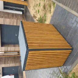 Nowoczesny śmietnik ogrodowy z drewnianymi listwami i metalową ramą, ustawiony na szarej kostce brukowej przed budynkiem z drewnopodobną elewacją i szarą bramą garażową.