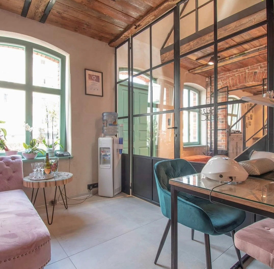 Loftowe wnętrze salonu kosmetycznego z metalową ścianką działową, drewnianym stropem i różową sofą. Widoczny stolik do manicure i dyspenser wody.