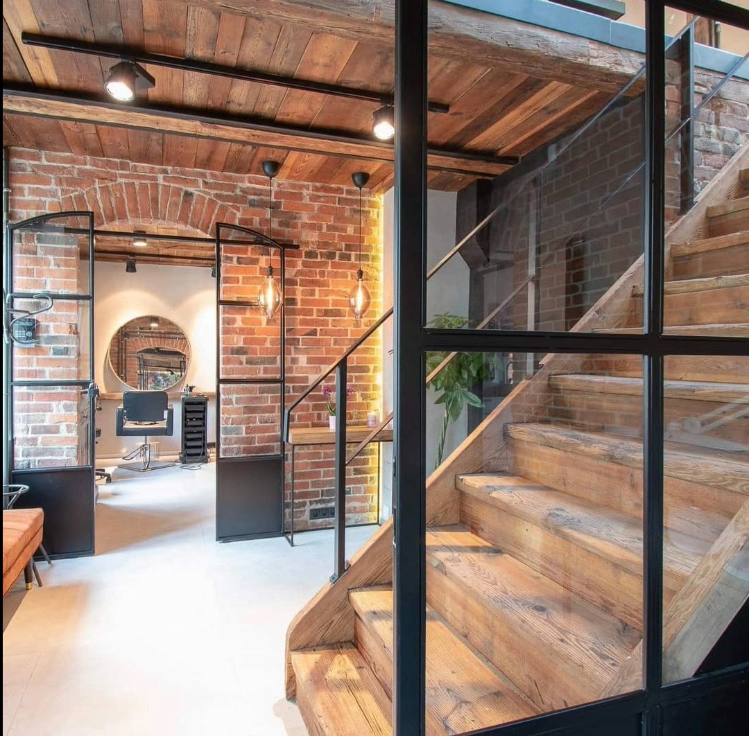Loftowe schody drewniane z metalową balustradą i szklaną ścianą działową, widok na ceglaną ścianę i otwarte drzwi do salonu fryzjerskiego. Industrialny design.