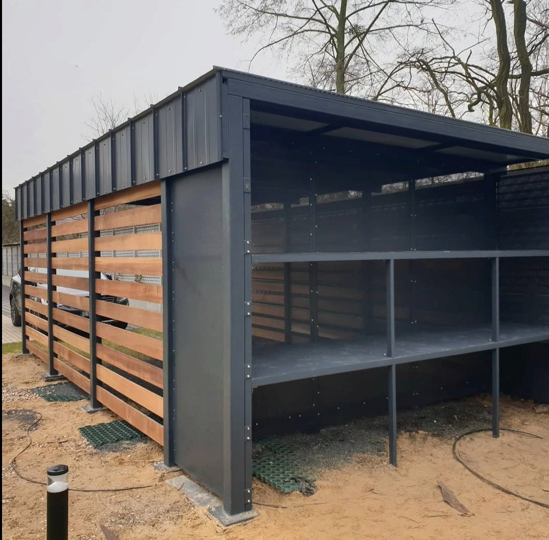 Nowoczesna konstrukcja z metalu i drewna: zadaszona wiata z drewnianym ogrodzeniem lamelowym, szara metalowa rama, widoczne półki wewnątrz, na zewnątrz kostka trawnikowa.