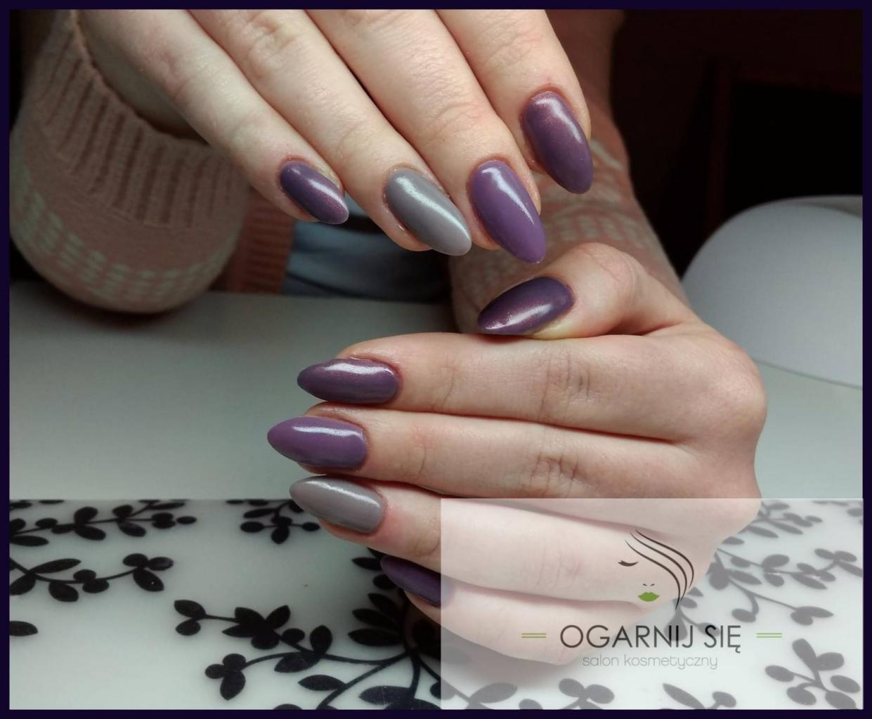 Dłonie z pomalowanymi paznokciami w odcieniach fioletu i szarości, na stole z motywem roślinnym. Na pierwszym planie logo salonu kosmetycznego.