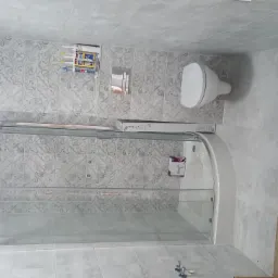 Wykończona łazienka z szarymi płytkami w ornamenty, kabiną prysznicową z przezroczystego szkła, podwieszaną toaletą i kratką wentylacyjną; widoczna kartonowa osłona na podłodze.