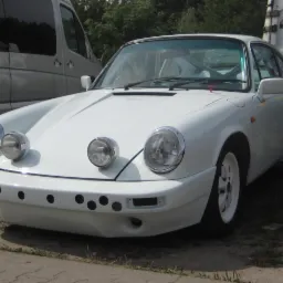 Białe Porsche 911 z dodatkowymi reflektorami i zmodyfikowanym zderzakiem z otworami, zaparkowane obok srebrnego vana i furgonetki z napisem 'Auto Blachy'.