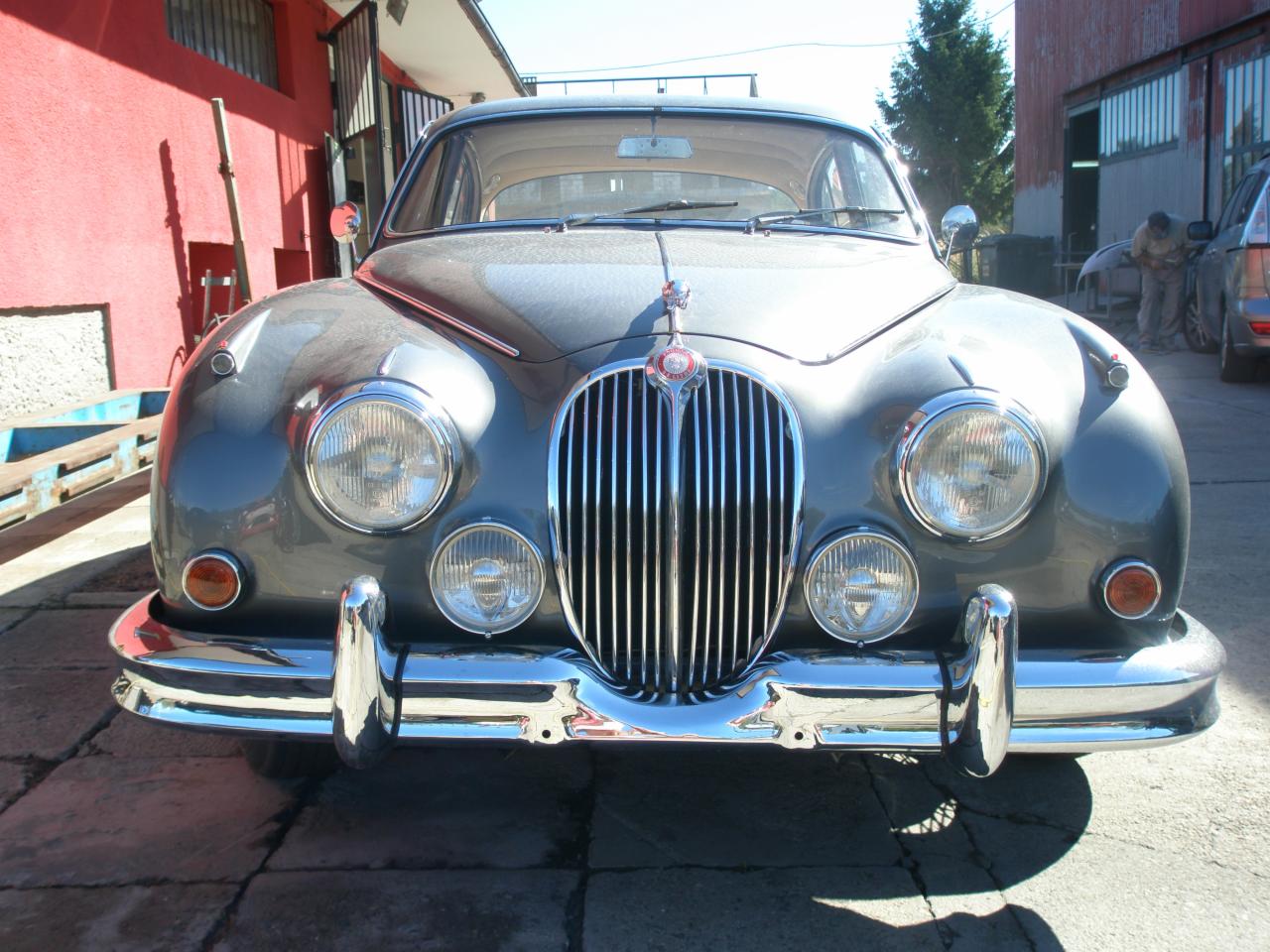 Przód klasycznego, szarego Jaguara z chromowanymi detalami, sfotografowany pod kątem, z widocznym warsztatem w tle.