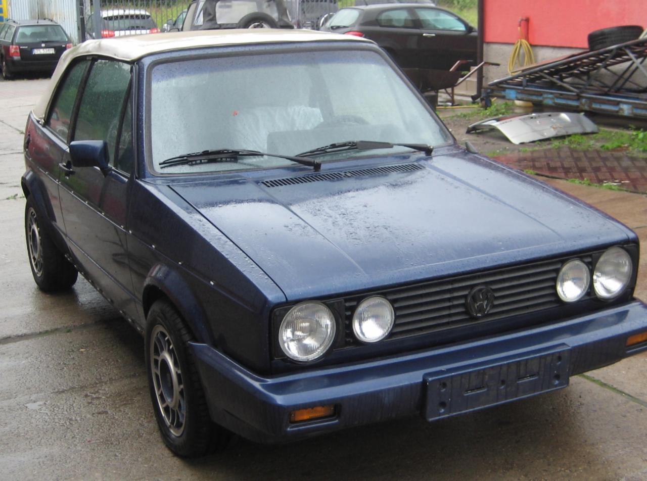 Granatowy Volkswagen Golf Cabriolet z jasnym dachem, zaparkowany na mokrym placu, widoczne krople deszczu na karoserii, w tle budynek i inne pojazdy.