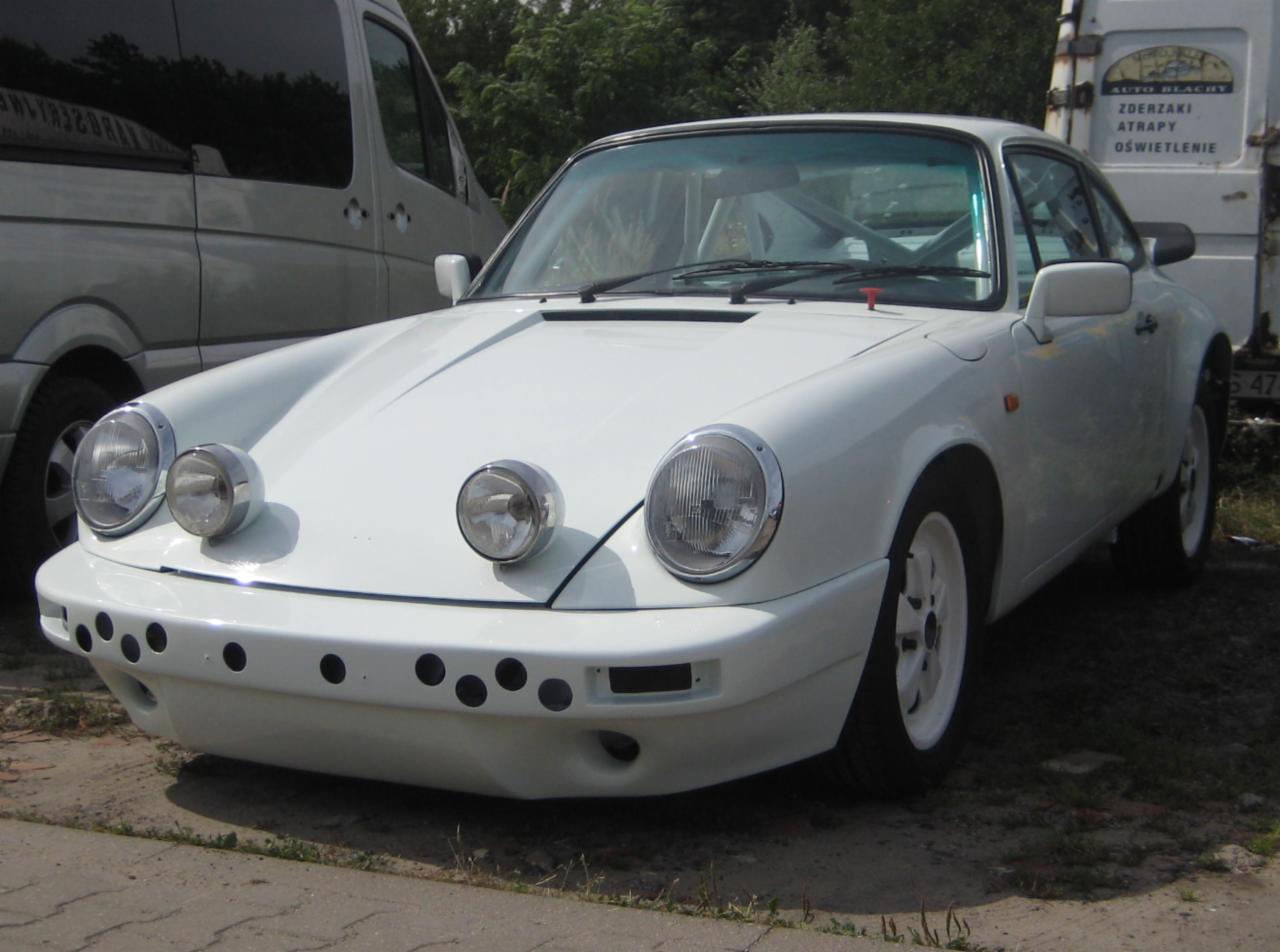 Białe Porsche 911 z dodatkowymi reflektorami i zmodyfikowanym zderzakiem z otworami, zaparkowane obok srebrnego vana i furgonetki z napisem 'Auto Blachy'.