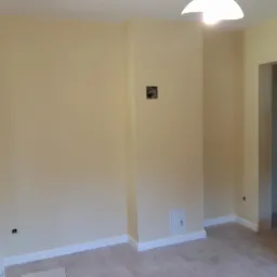Pokój po remoncie: ściany pomalowane na jasnożółty kolor, białe listwy przypodłogowe, beżowe płytki podłogowe, widoczne puszki elektryczne i otwór wentylacyjny.