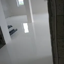 Świeżo wylana, jasnoszara, błyszcząca podłoga w surowym wnętrzu, widoczne narzędzia i fragment okna w oddali.