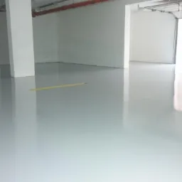 Jasnoszara, błyszcząca podłoga w pustym pomieszczeniu przemysłowym z białymi ścianami i słupem, widoczne rury pod sufitem i otwarta brama garażowa.