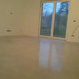 Świeżo wylana, gładka, jasnoszara posadzka w surowym wnętrzu z widocznymi puszkami elektrycznymi i przewodami na ścianie, duże okno balkonowe z widokiem na drzewa.