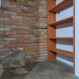 Ręcznie robiona, drewniana półka wnękowa na tle ściany z cegły i kamiennej podłogi. Widoczne słoje drewna i unikalna faktura cegieł. Surowy, industrialny styl.