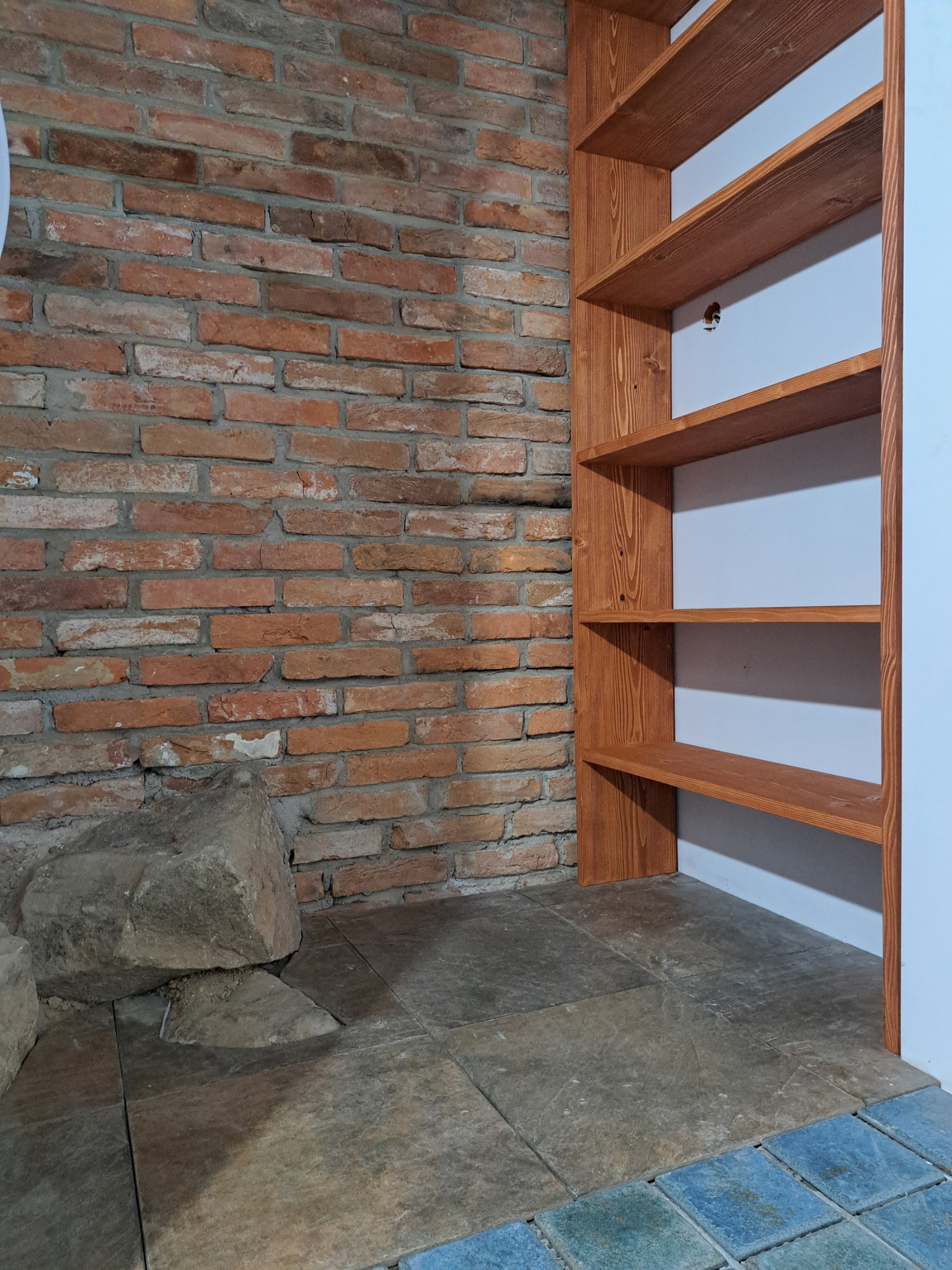 Ręcznie robiona, drewniana półka wnękowa na tle ściany z cegły i kamiennej podłogi. Widoczne słoje drewna i unikalna faktura cegieł. Surowy, industrialny styl.