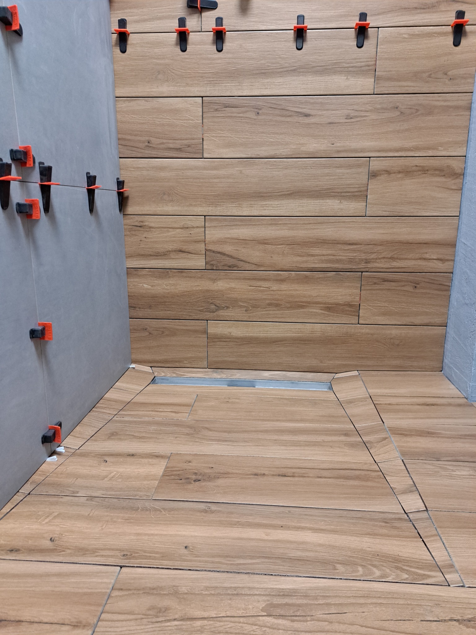 Układanie płytek imitujących drewno na ścianie i podłodze. Widoczne klipsy poziomujące, odpływ liniowy. Płytki ułożone poziomo, z przesunięciem, tworząc efekt drewnianej podłogi.