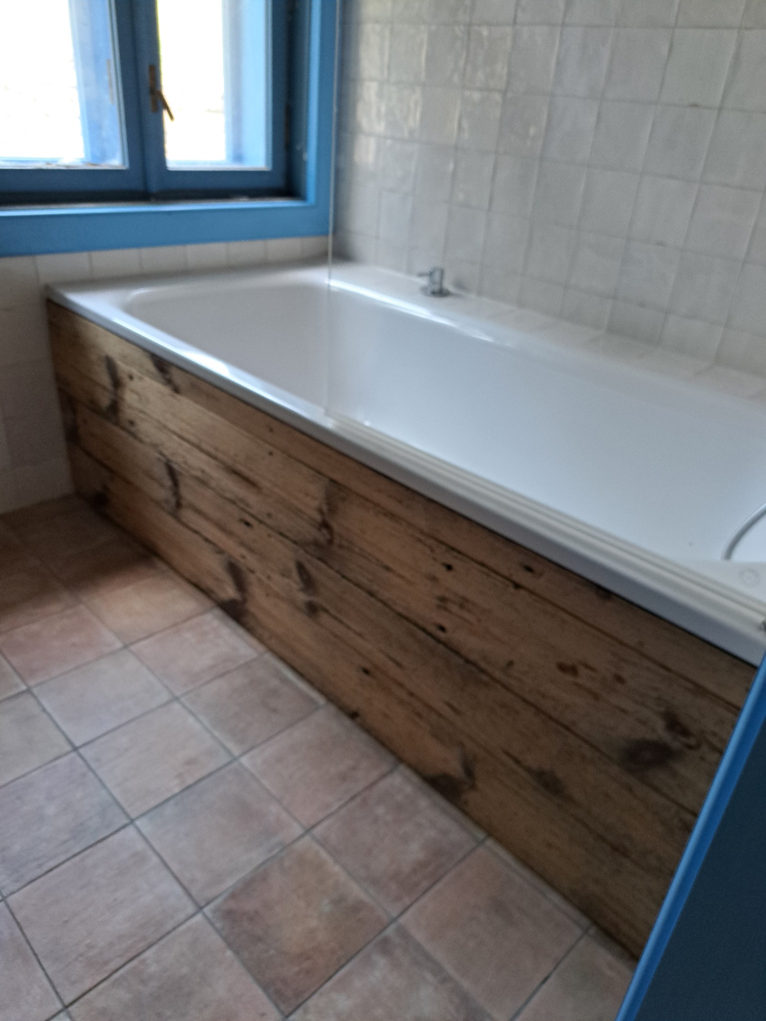 Biała wanna z drewnianą obudową w łazience w Laskowej. Okno z niebieską ramą i kafelki na ścianie. Podłoga wyłożona jest terakotą w kwadraty.