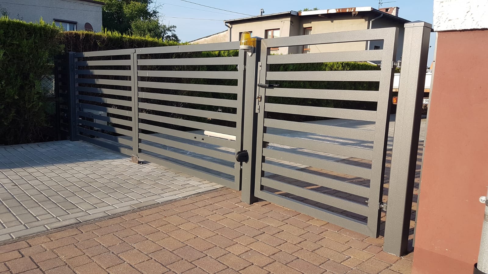 Nowoczesna, grafitowa brama wjazdowa z poziomych profili, częściowo otwarta, z widocznym zamkiem i sygnalizatorem świetlnym, na tle kostki brukowej.