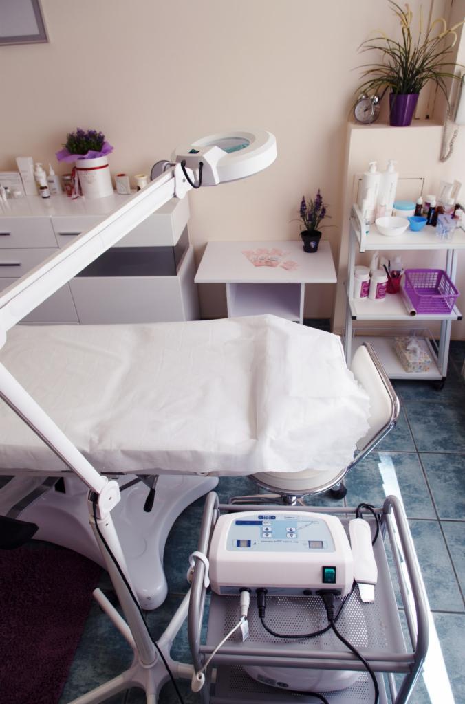 Przygotowane stanowisko zabiegowe z lampą powiększającą, urządzeniem kosmetycznym na wózku i półkami z kosmetykami w tle.