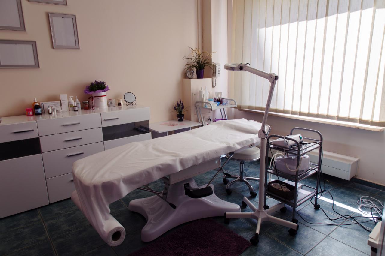 Jasne wnętrze gabinetu z łóżkiem kosmetycznym przykrytym białym prześcieradłem, lampą powiększającą i wózkiem z urządzeniami kosmetycznymi, na ścianie wiszą puste ramki.