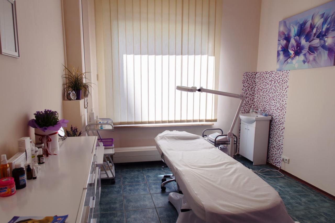 Jasne wnętrze gabinetu z łóżkiem kosmetycznym przykrytym białym prześcieradłem, lampą zabiegową, szafką z umywalką, oraz komodą z kosmetykami i kwiatami w doniczce.