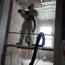 Pracownik w kombinezonie ochronnym na rusztowaniu aplikuje pianę izolacyjną na sufit w surowym wnętrzu budynku z odsłoniętą cegłą.