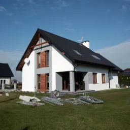 Nowoczesny dom jednorodzinny z białymi ścianami i szarymi akcentami, czarny dach z oknami dachowymi, widoczne rusztowania i materiały na trawniku, sugerujące prace wykończeniowe lub remontowe...