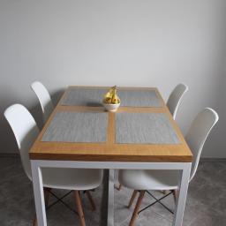 Beleco - Stół z drewnianym blatem i białymi, metalowymi nogami, otoczony czterema białymi krzesłami. Na stole leżą szare podkładki i stoi miseczka z bananami.