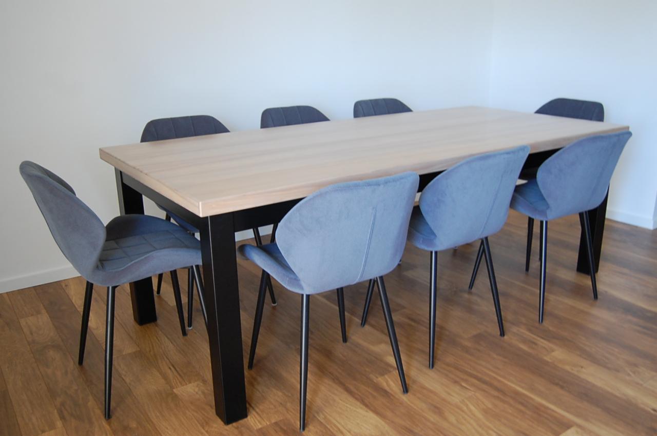 Stół z jasnym drewnianym blatem i czarnymi nogami otoczony sześcioma krzesłami z szarobłękitną tapicerką i czarnymi, smukłymi nogami, ustawiony na brązowej podłodze.