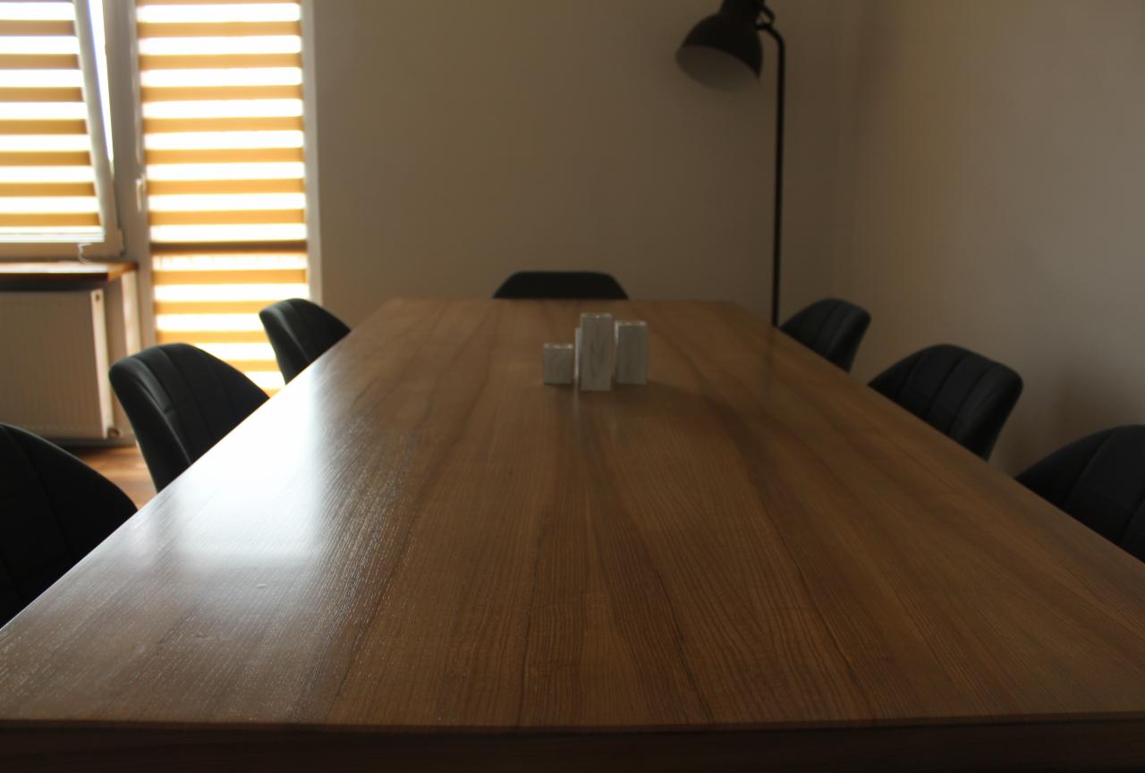 Długi, drewniany stół z krzesłami i minimalistyczną dekoracją z trzech białych, prostopadłościennych brył, oświetlony lampą stojącą, ustawiony na tle okna z żaluzjami.