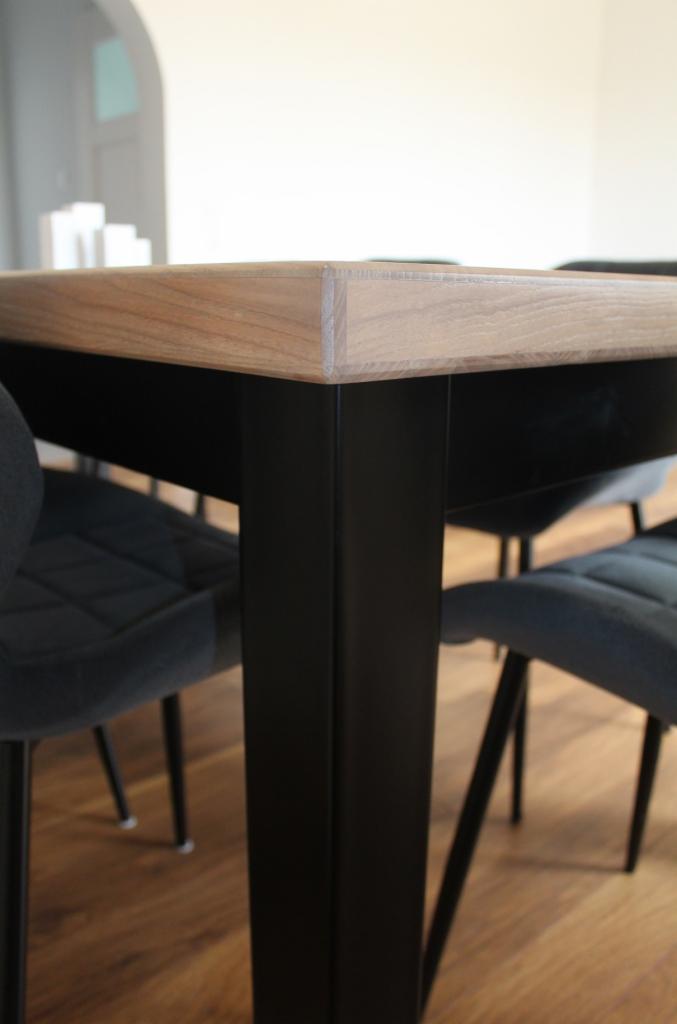 Fragment stołu z blatem z jasnego drewna i czarnymi, metalowymi nogami, widoczne krzesła z ciemną tapicerką w tle, parkiet na podłodze.