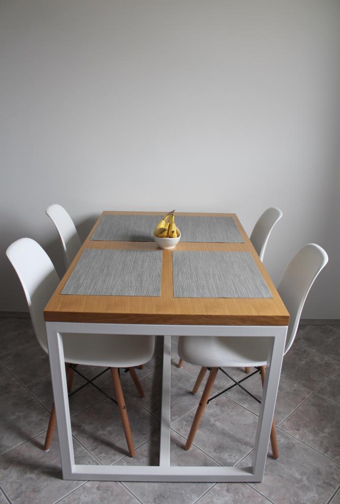 Stół z drewnianym blatem i białymi, metalowymi nogami, otoczony czterema białymi krzesłami. Na stole leżą szare podkładki i stoi miseczka z bananami.