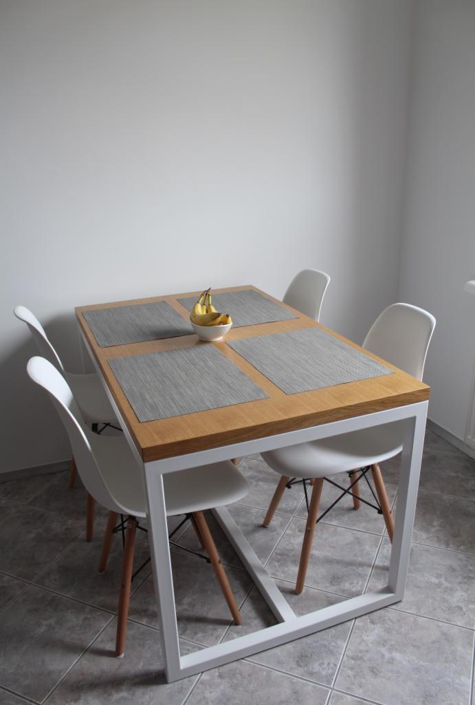 Stół z drewnianym blatem i białą, metalową podstawą w kształcie litery Z, nakryty szarymi podkładkami i miską z bananami, otoczony czterema białymi krzesłami na drewnianych nogach.