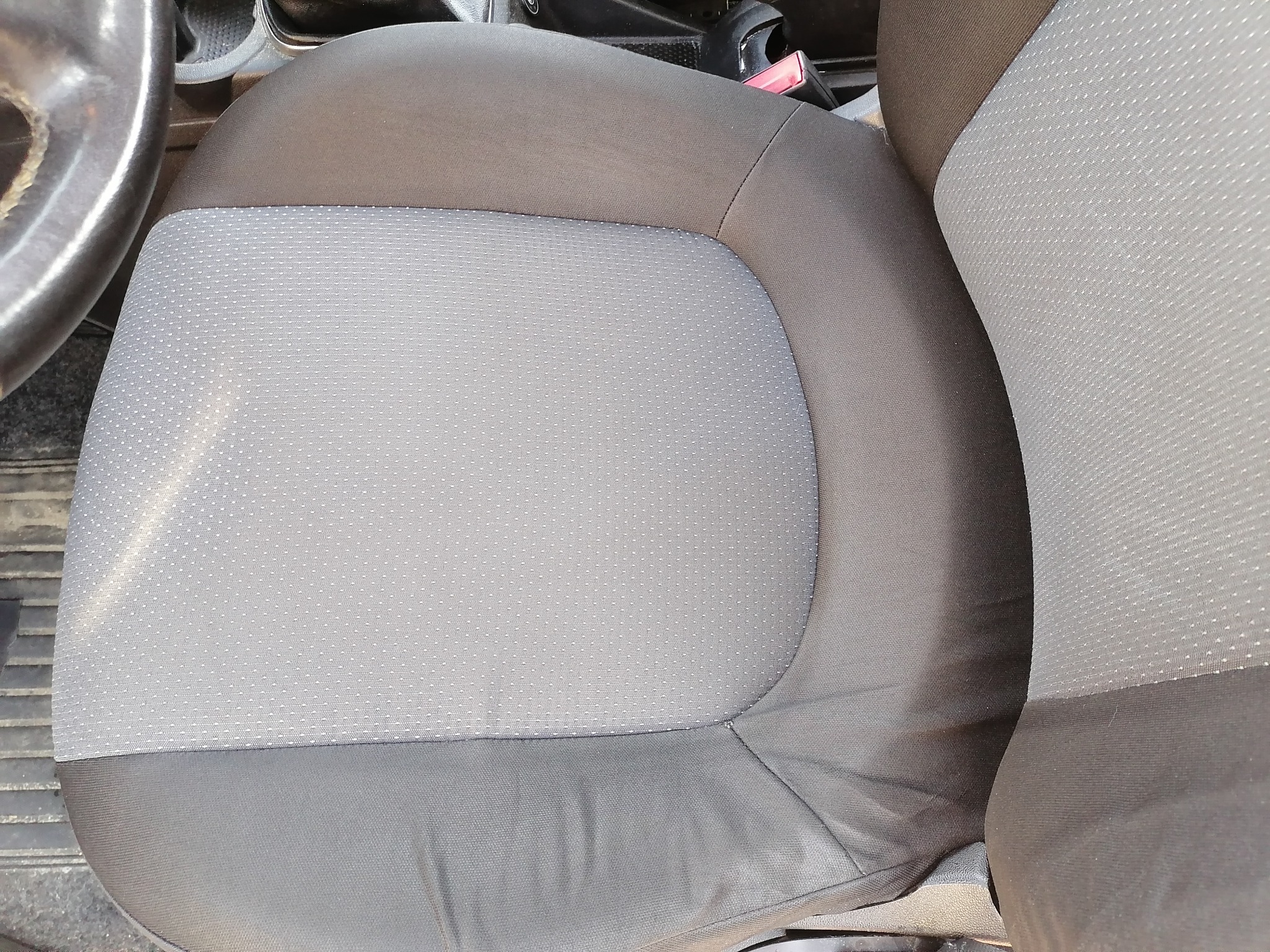 Szare, materiałowe siedzenie samochodowe z widoczną kierownicą i pasem bezpieczeństwa.