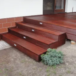 Drewniany taras z egzotycznego drewna bangkirai, z trzema stopniami schodów wyposażonymi w oświetlenie punktowe, przylegający do budynku z ceglaną podmurówką i fragmentem trawnika z małą rośliną...