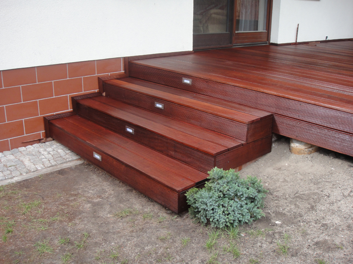 Drewniany taras z egzotycznego drewna bangkirai, z trzema stopniami schodów wyposażonymi w oświetlenie punktowe, przylegający do budynku z ceglaną podmurówką i fragmentem trawnika z małą rośliną...