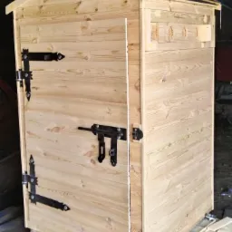 Surowa, drewniana wędzarnia z czarnymi okuciami, umieszczona na palecie, widoczne otwory wentylacyjne i proste zapięcie, tło warsztatowe.